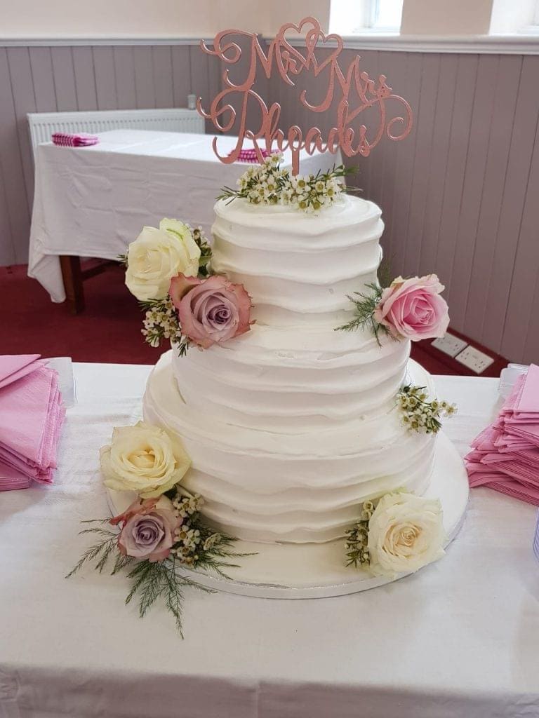 Three-tier white wedding cake decorated with roses and greenery, topped with a "Mr & Mrs Rhoades" sign. Pink serviettes are stacked nearby on the table.