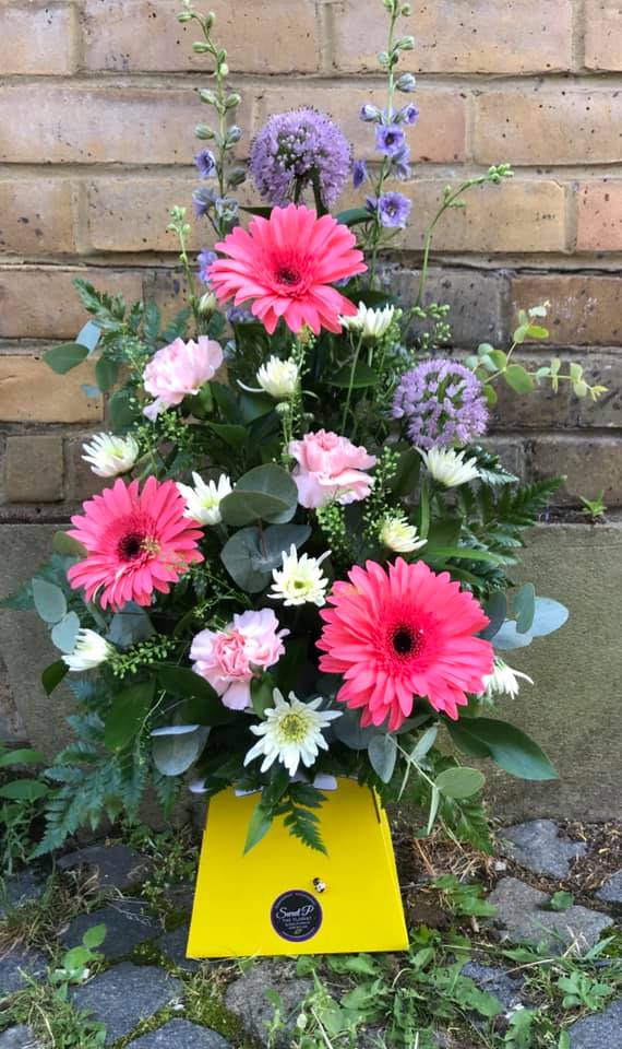 Jeoffrey is a floral arrangement featuring pink gerbera daisies, assorted white and pink blooms, and greenery in a yellow box, displayed against a brick wall.