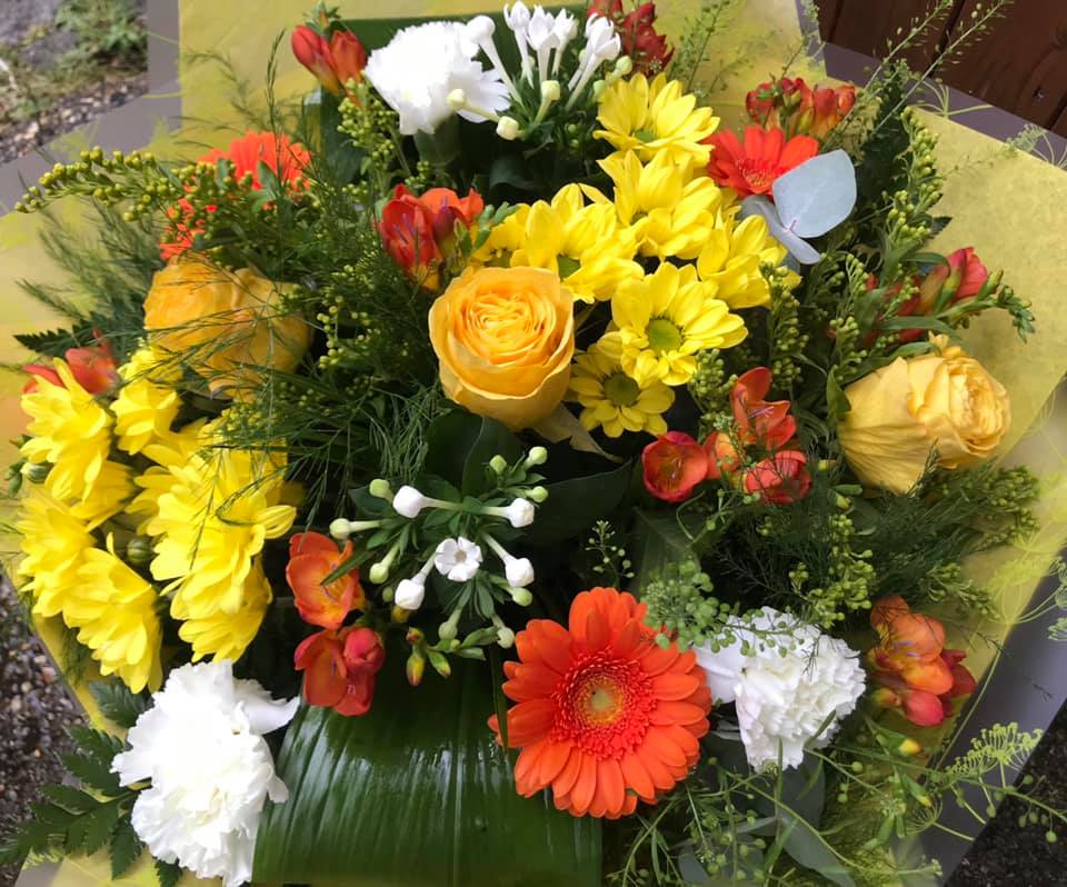 A bouquet of yellow roses, yellow daisies, orange and red gerbera daisies, white carnations, and green foliage arranged together.