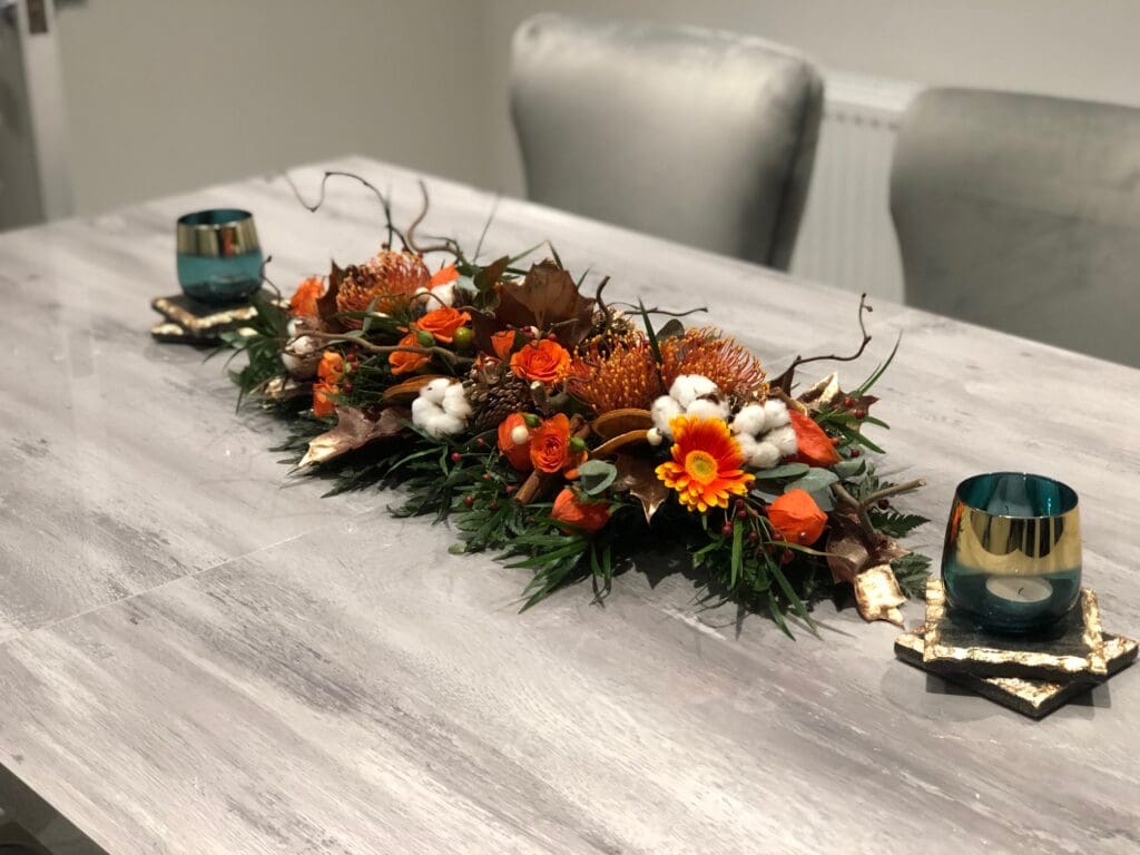 A rectangular dining table with a floral centrepiece of orange, yellow, and white flowers, green foliage, and two tealight candles in blue holders on either side.