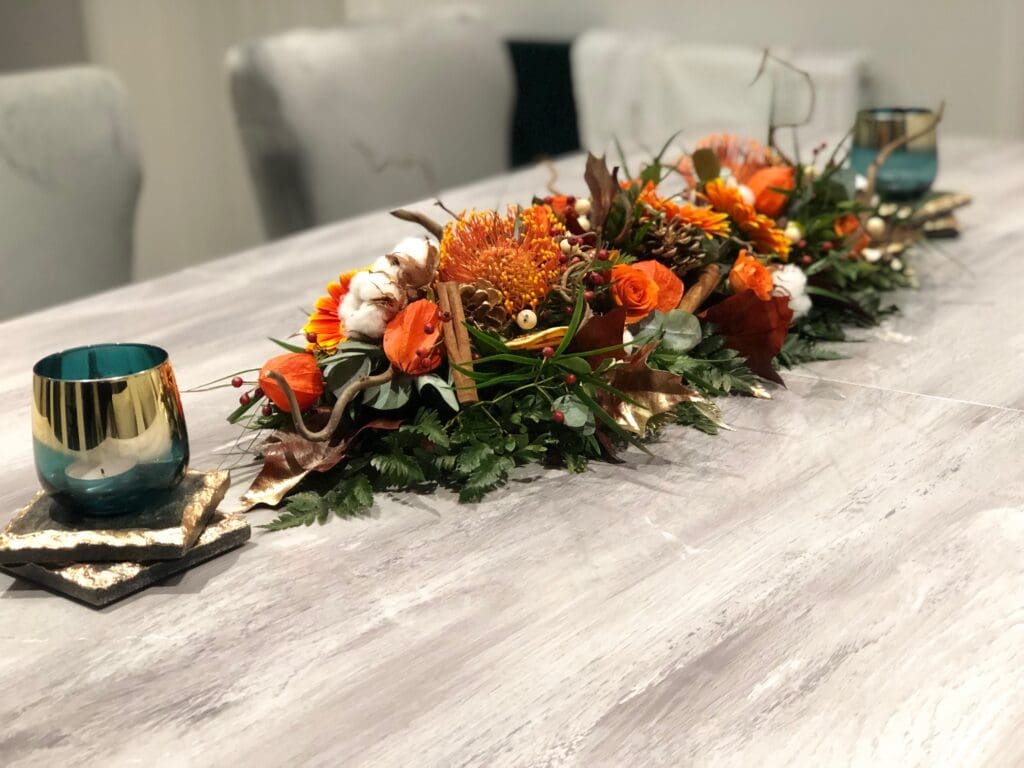 A floral centrepiece with orange and white flowers, greenery, and decorative gold elements sits on a light-coloured dining table with two glass candle holders.