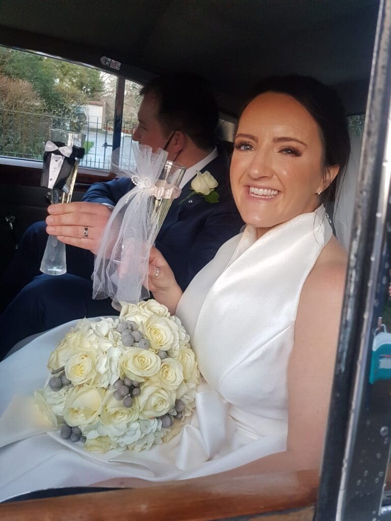 A bride in a white dress holds a bouquet of white roses and a decorated glass, smiling whilst sitting next to a man in a suit inside a car.