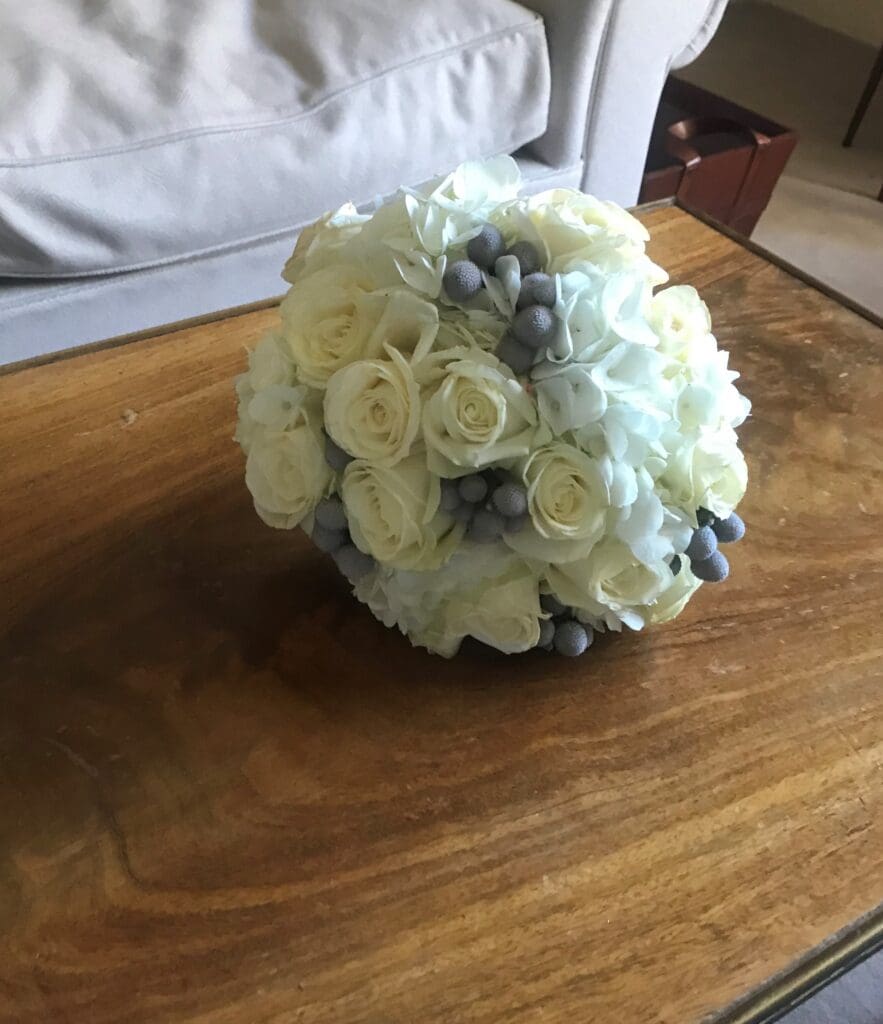 A bouquet of white roses, hydrangeas, and grey berries sits on a wooden table in front of a beige sofa.