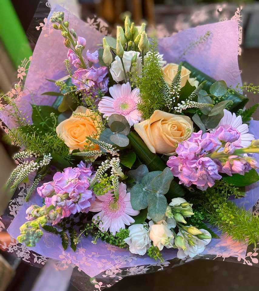 A bouquet of yellow roses, pink gerbera daisies, and purple flowers with greenery, wrapped in pink decorative paper.