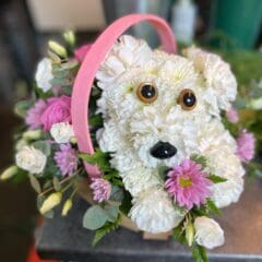 Floral basket arrangement shaped like a white dog, featuring white carnations, pink roses, pink chrysanthemums, eucalyptus foliage, and greenery in a pink handled basket.