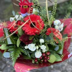 Festive Christmas flower bouquet with red gerbera daisies, white roses, green foliage, red berries, and a small Santa decoration wrapped in red paper.