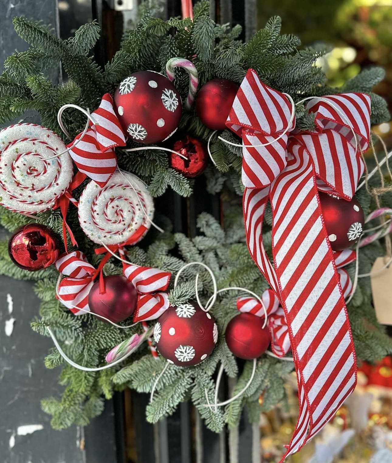 Festive Christmas door wreath made with fresh fir foliage, red baubles, candy canes, peppermint swirl decorations, and large red-and-white striped ribbon bows for a candy cane themed design.