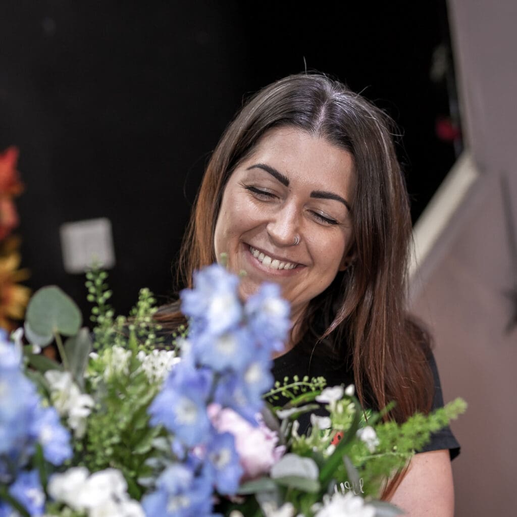 Smiling florist with long brown hair arranging fresh flowers, surrounded by soft blue delphiniums, white blooms, eucalyptus, and greenery in a flower shop setting.